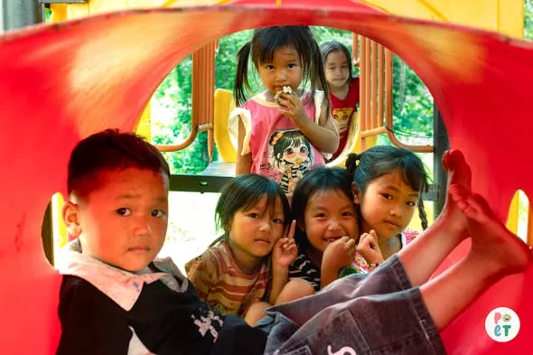 Các em nhỏ ngồi trong ống chui của cầu trượt, cười nhìn vào ống kính.