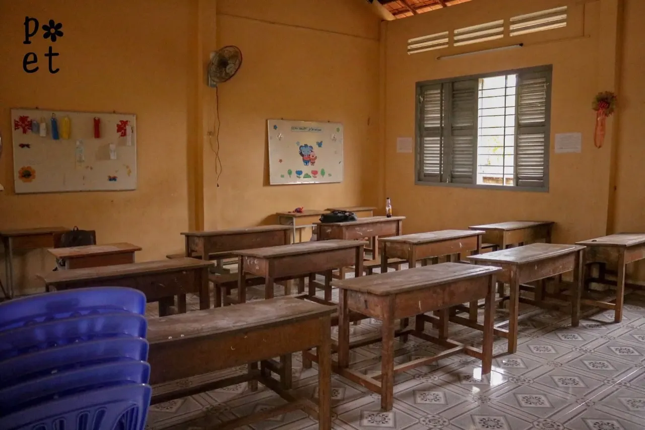 Lớp học tại điểm trường với bàn ghế gỗ đã cũ, quạt treo tường và cửa sổ chớp